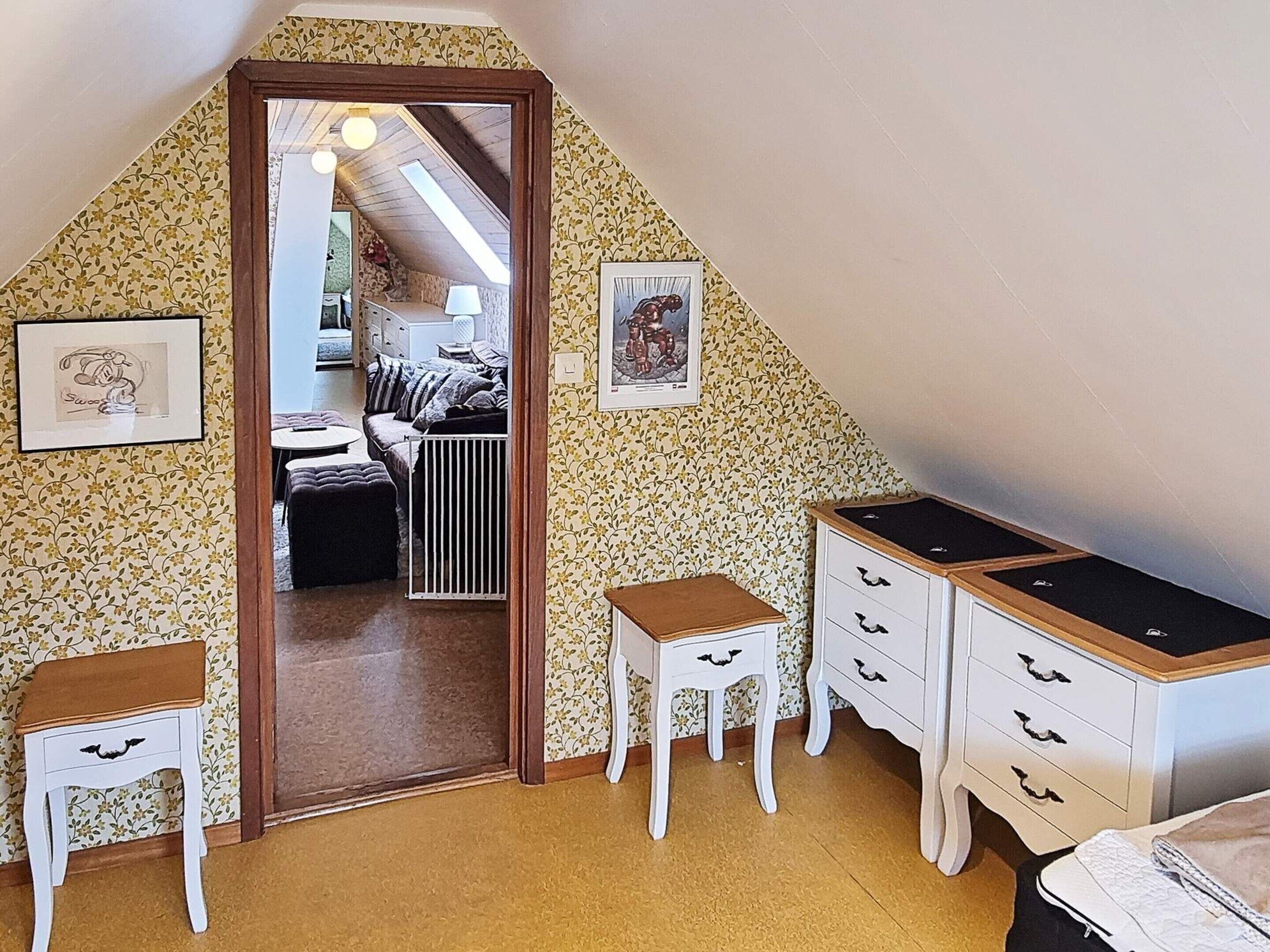 Un dormitorio con techo inclinado, una cama, un aparador, dos sillas y un radiador.