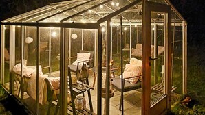 Een glazen kas met houten frame, waarin diverse stoelen en tafels staan, en een klein tafeltje met een kaars.