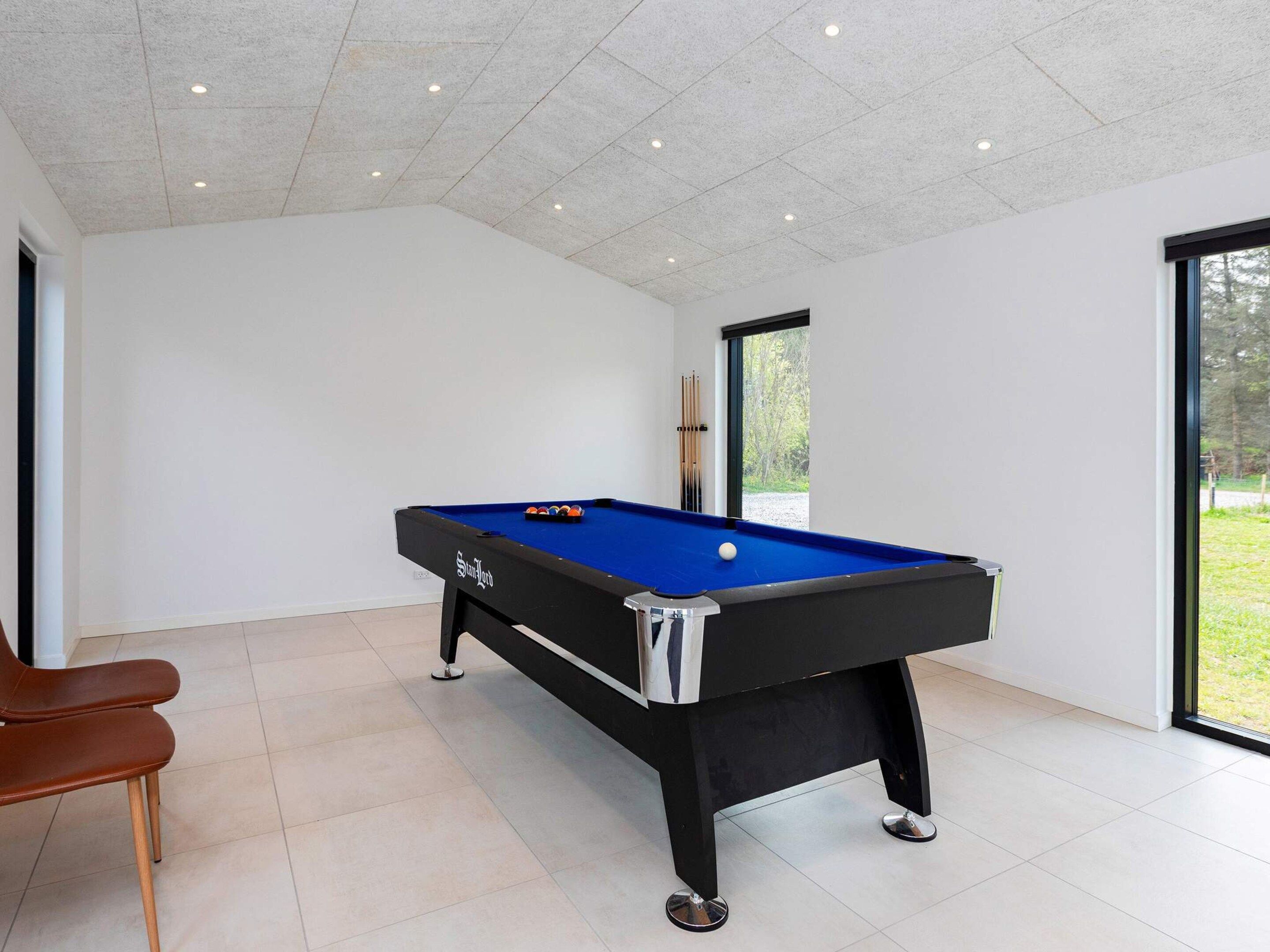 A pool table in a room with white walls, a brown chair, and a window providing a view of greenery outside.