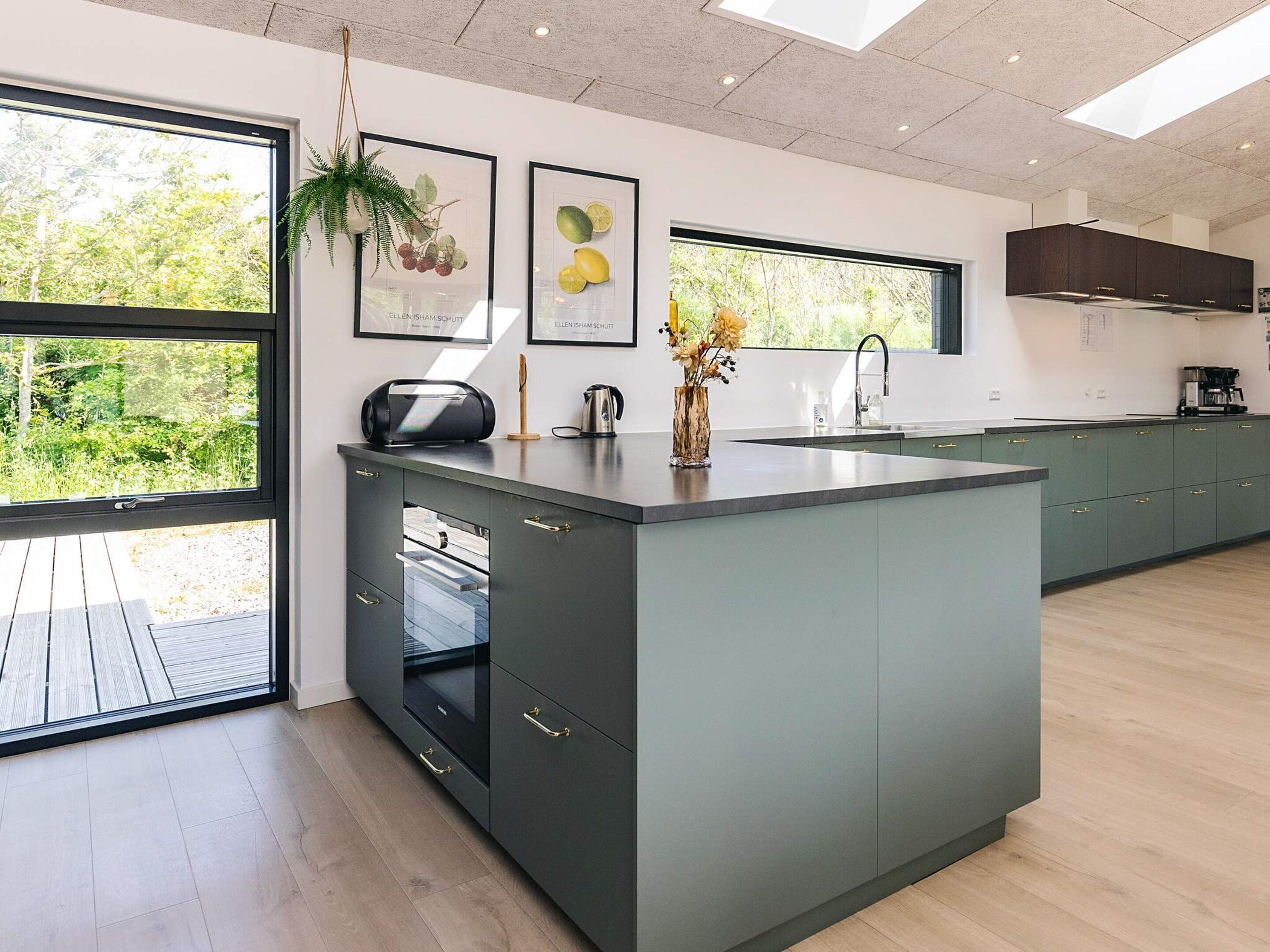 A modern kitchen with a large island, dark cabinets, and a view of greenery outside.