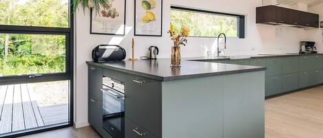 A modern kitchen with a large island, dark cabinets, and a view of greenery outside.