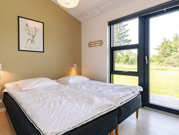 Une chambre à coucher avec un lit, des lampes de chevet, un tableau encadré et une grande fenêtre donnant sur un paysage verdoyant.
