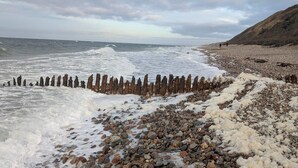 A rocky beach with waves crashing onto the shore, a wooden fence, and a hilly landscape in the background. - 6 Person Holiday Home in Tisvildeleje (Tisvildeleje)