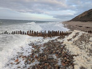 Ein felsiger Strand mit brechenden Wellen, eine Holzlattenzäunung und eine hügelige Landschaft im Hintergrund.