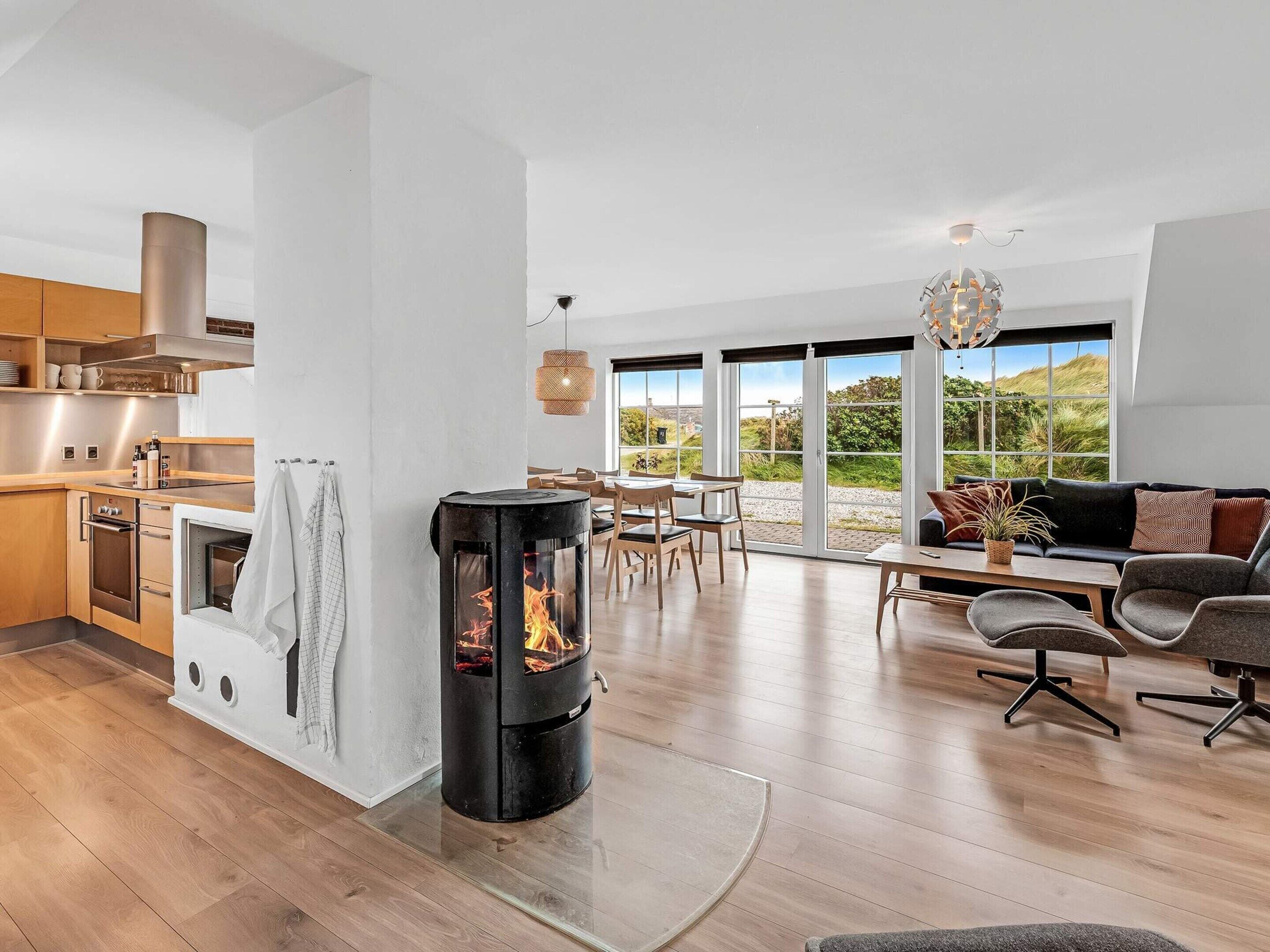 A modern kitchen with a wood-burning stove, a dining area with a view, and a cozy living space with a sofa and a chandelier.