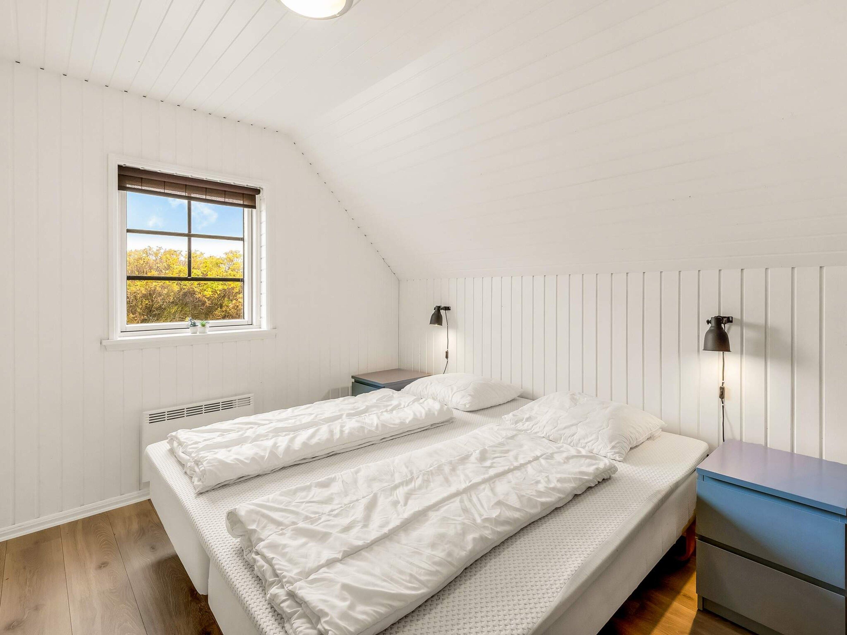 A bedroom with a sloped ceiling, a window with blinds, a bed with white bedding, two bedside lamps, and a nightstand with a lamp.