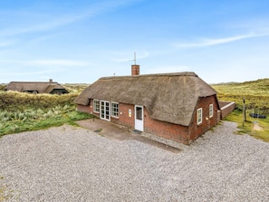 A thatched-roof cottage with a gravel driveway, surrounded by grassy fields and hills. - 5 Star Holiday Home in Ringkobing (Ringkobing)