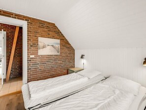A bedroom with a sloped ceiling, a brick wall, a bed with white bedding, a wooden door, and a picture of a snowy landscape. - 5 Star Holiday Home in Ringkobing (Ringkobing)