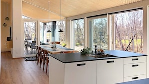 A modern kitchen with a large island, wooden flooring, and large windows offering views of the outdoors.