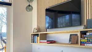 A modern living room with a large flat-screen TV mounted on a wooden wall, a bookshelf with various books and board games, and two hanging pendant lights.