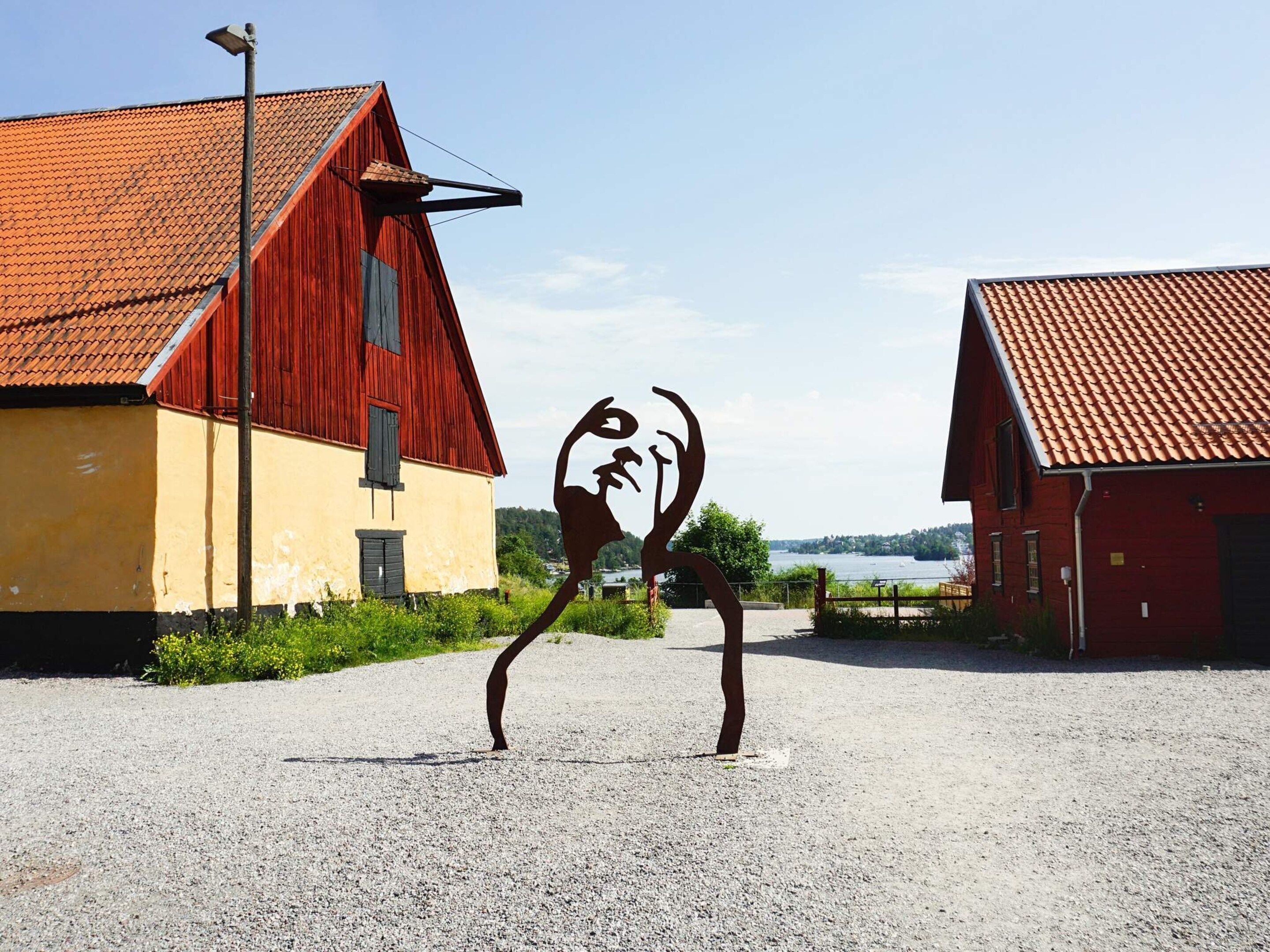 A sculpture in a gravel area with two red barns in the background.