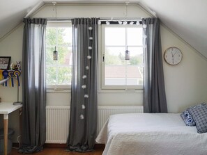 A bedroom with a bed, a window with curtains, a radiator, a wall clock, and a desk with a lamp.