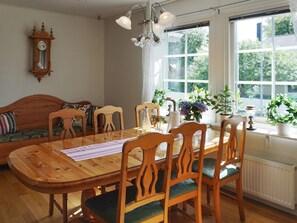 A dining area with a wooden table and chairs, a clock on the wall, and a window with plants.