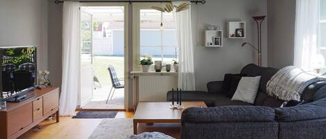 A living room with a sofa, a coffee table, a TV on a cabinet, and a view through a door to an outdoor area.