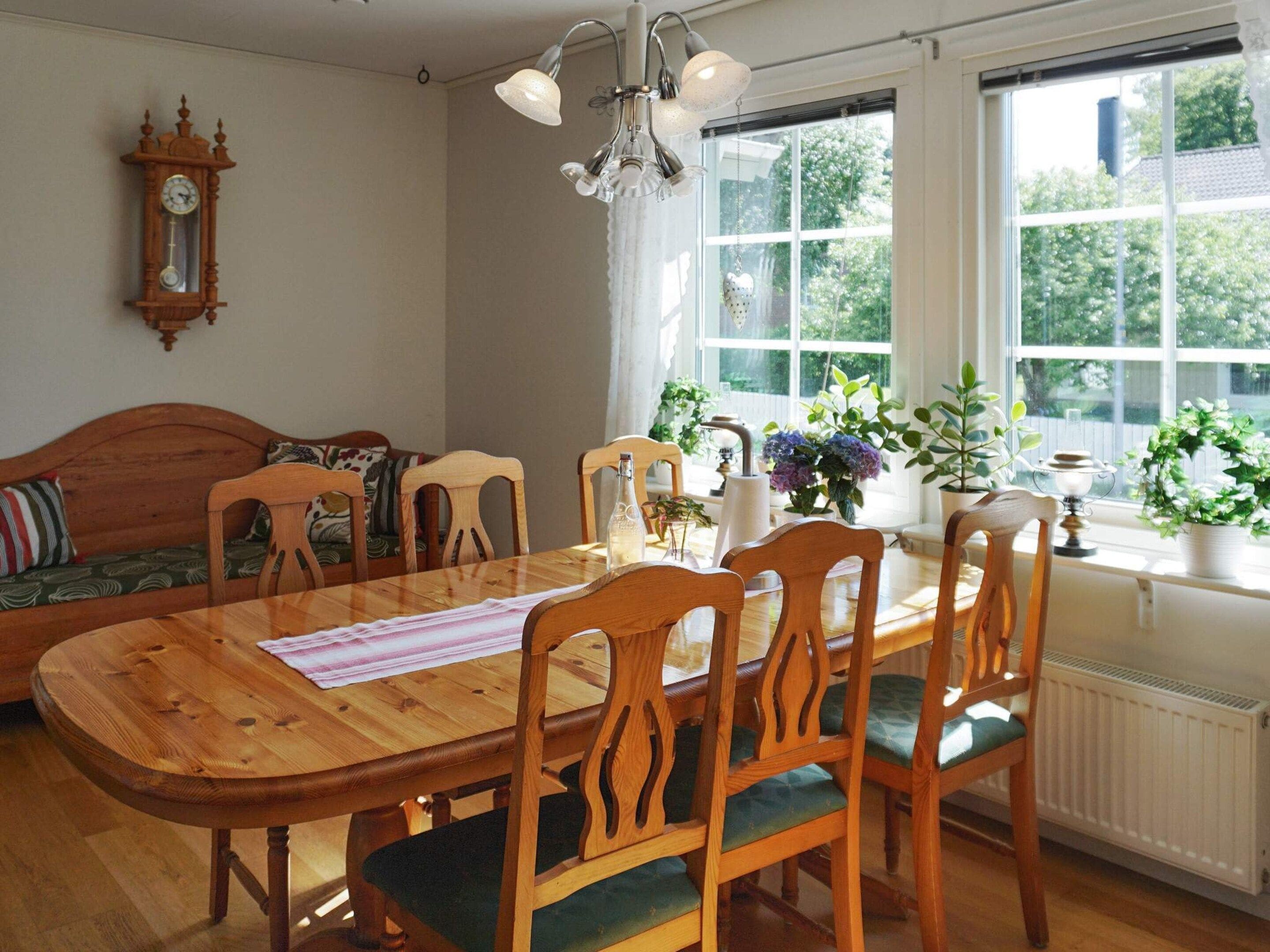 En matplats med ett träbord och stolar, en klocka på väggen och ett fönster med växter.