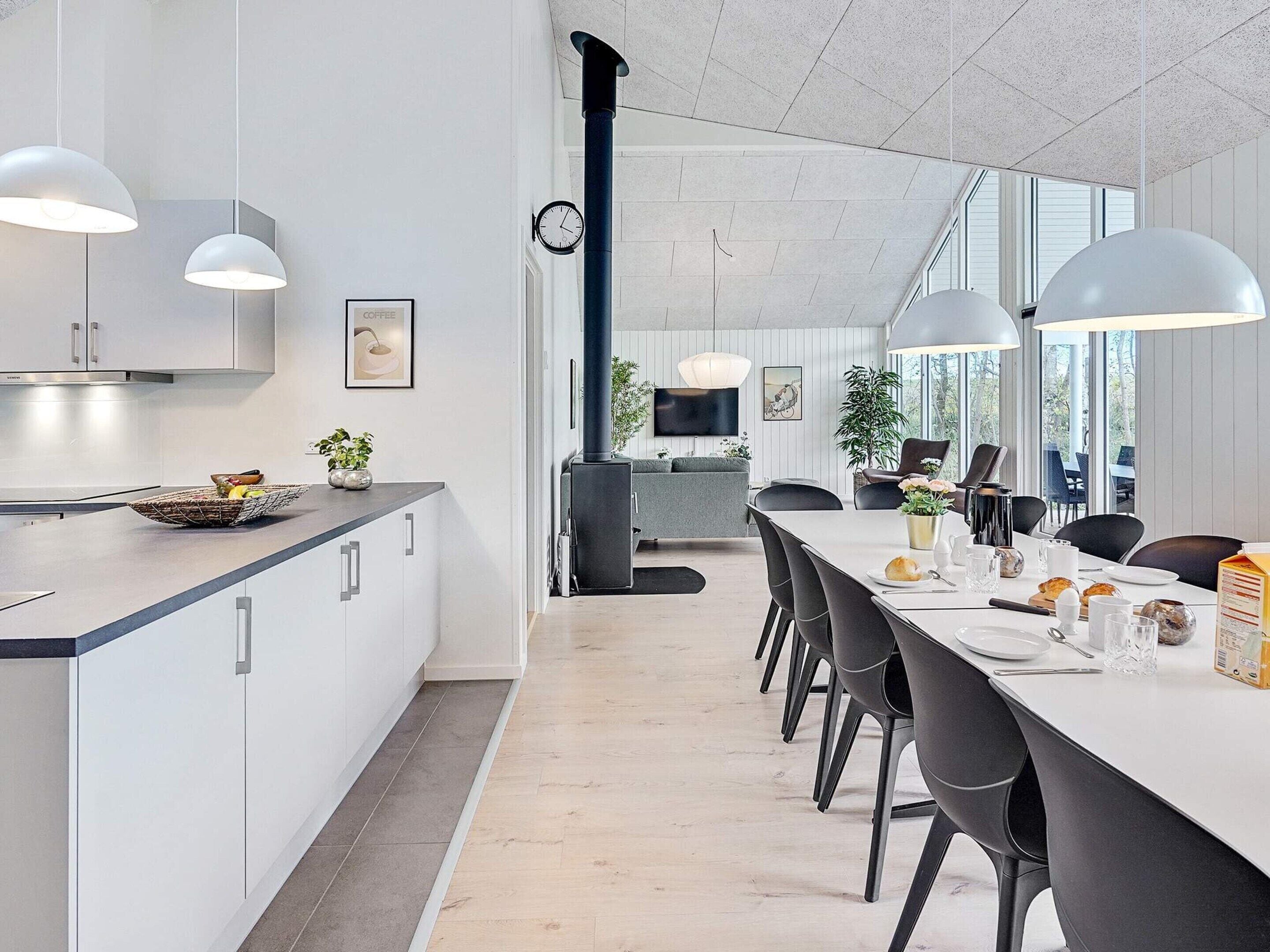 Ett modernt kök med vita skåp, en matplats med ett långt bord och stolar samt ett stort fönster som släpper in naturligt ljus.