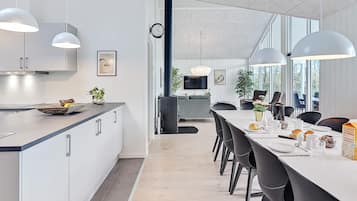 Ett modernt kök med vita skåp, en matplats med ett långt bord och stolar samt ett stort fönster som släpper in naturligt ljus.