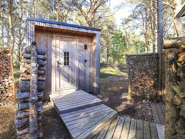 A wooden cabin with a door and a small window, surrounded by trees and a stone wall.