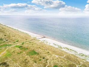 Vue aérienne d’un paysage côtier avec une plage de sable, des dunes herbeuses et un ciel bleu dégagé.