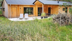 A modern wooden cabin with a large sliding glass door, a patio with two lounge chairs, and a surrounding natural landscape. - 5 Star Holiday Home in Albaek (Aalbaek)
