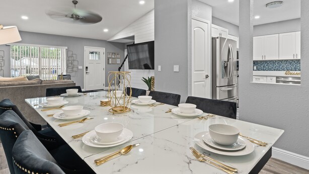 A modern dining room with a marble table set for six, a black leather couch, and a kitchen with white cabinets and a blue backsplash.