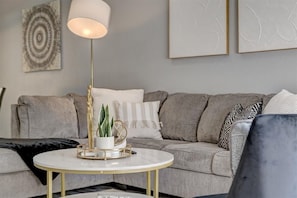 Een moderne woonkamer met een grijze bank, een ronde salontafel met gouden poten, een vloerlamp en abstracte muurkunst.