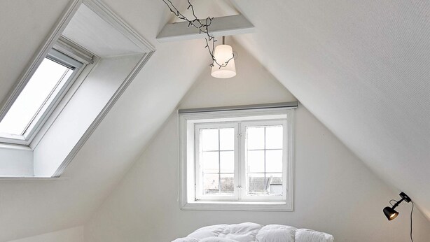 Une chambre avec un plafond en pente, un puits de lumière, une fenêtre, un lit avec une literie blanche et une lampe de chevet.