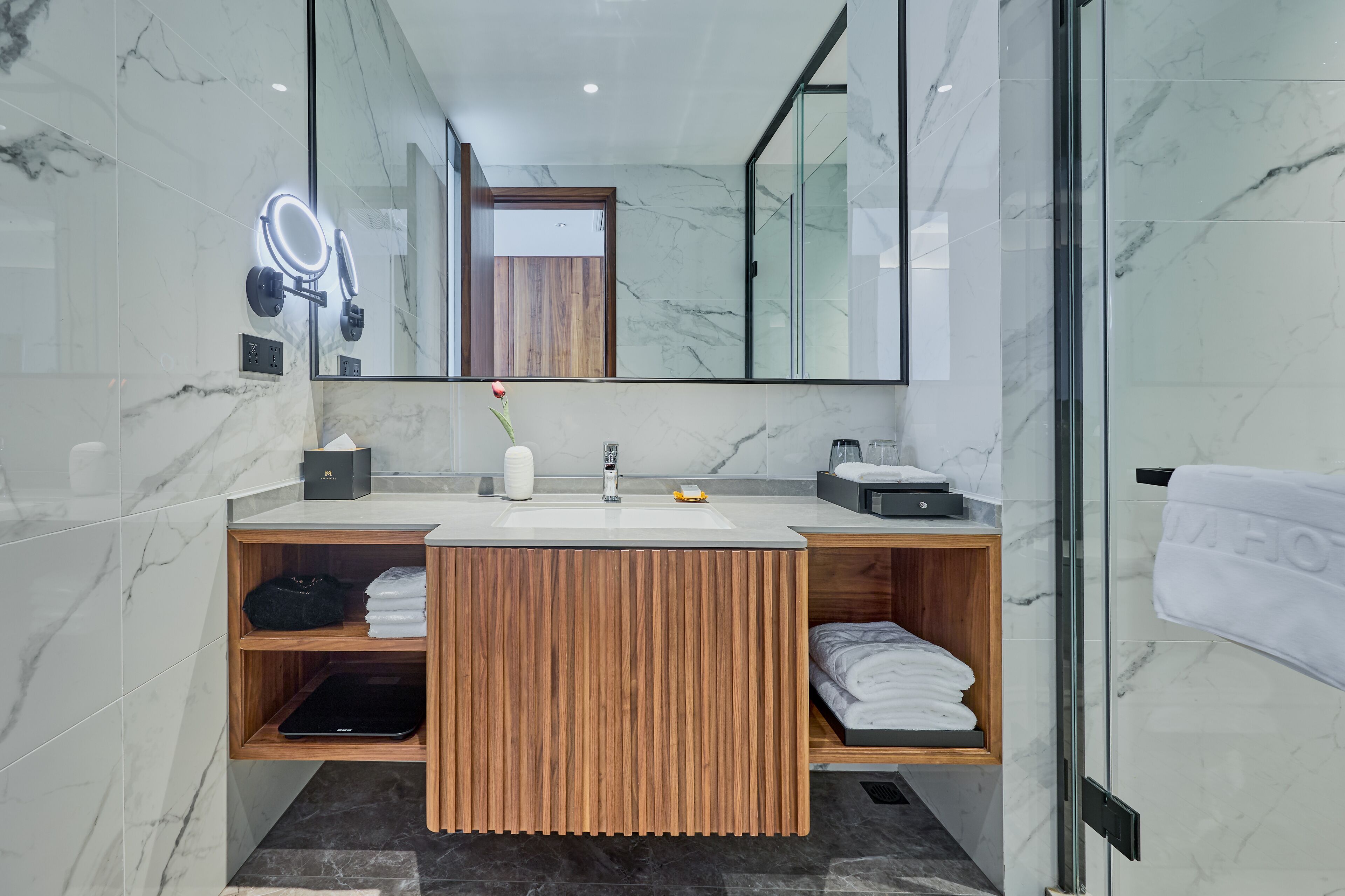 Ein modernes Badezimmer mit einem Holzkosmetikschrank, Marmorfliesen, einem großen Spiegel mit Beleuchtung und einer Glasdusche.