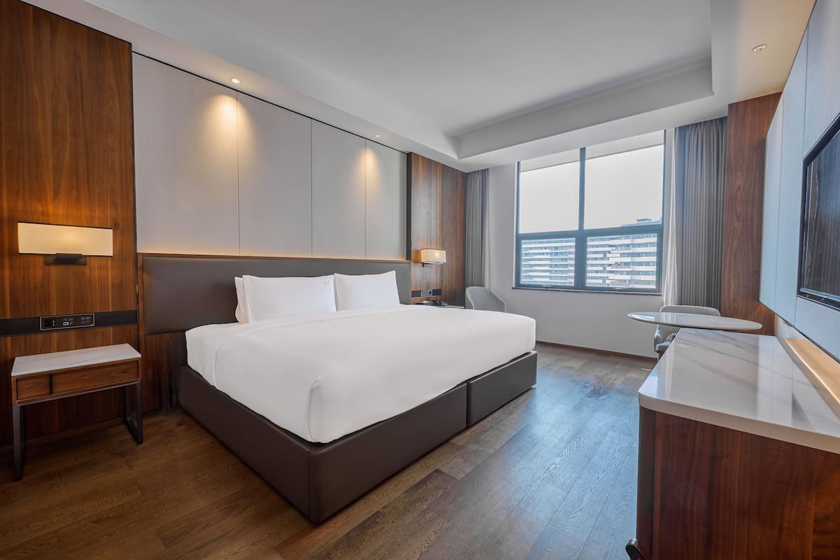 Ein modernes Hotelzimmer mit einem großen Bett, einem Nachttisch, einem Schreibtisch, einem Sessel, einem kleinen Tisch und einem großen Fenster mit Blick auf die Stadt.