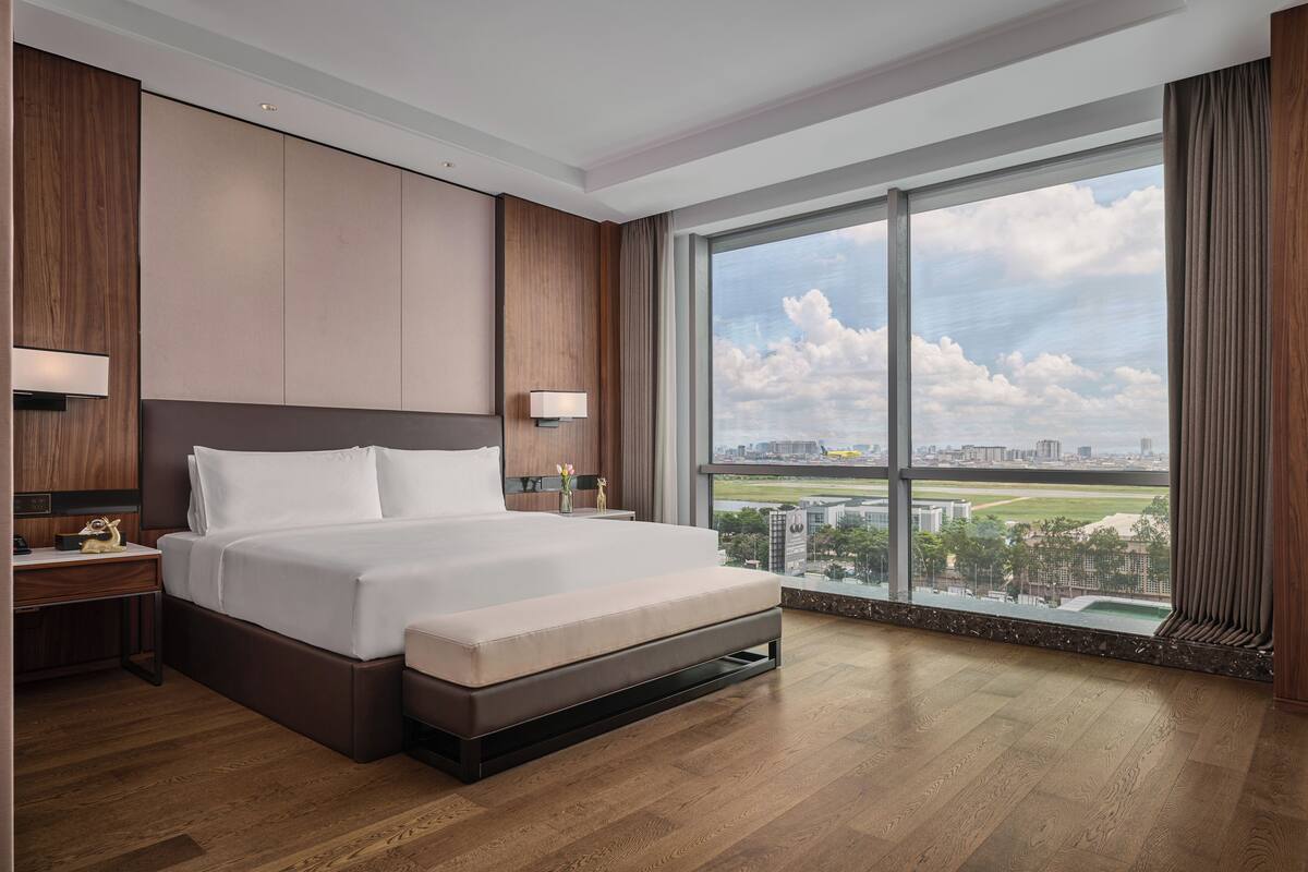 Ein modernes Hotelzimmer mit einem großen Bett, Holzboden, einem Nachttisch, einer Lampe und einem großen Fenster mit Blick auf die Stadt.