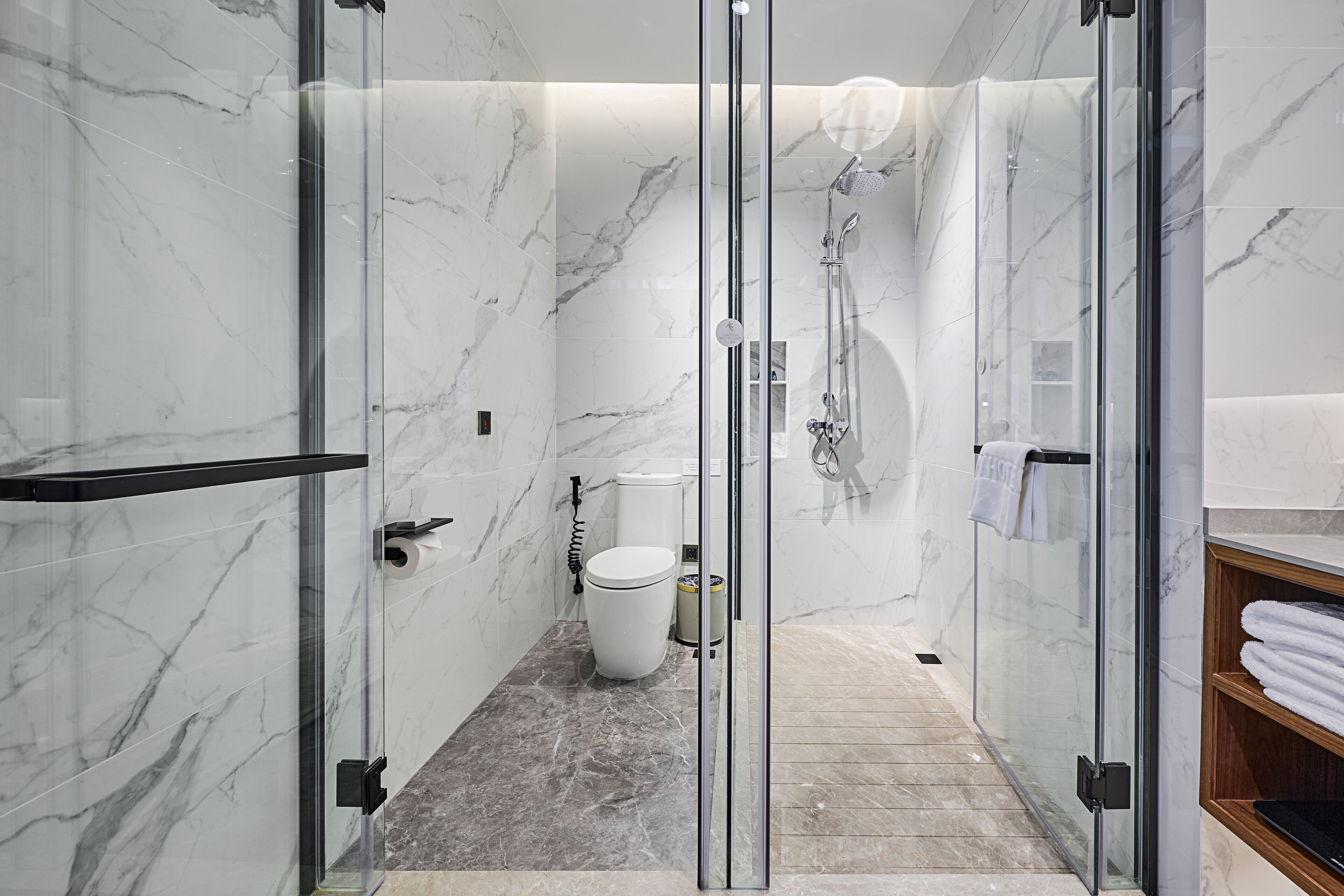 A modern bathroom with a glass-enclosed shower, marble walls, and a wooden shelf with towels.