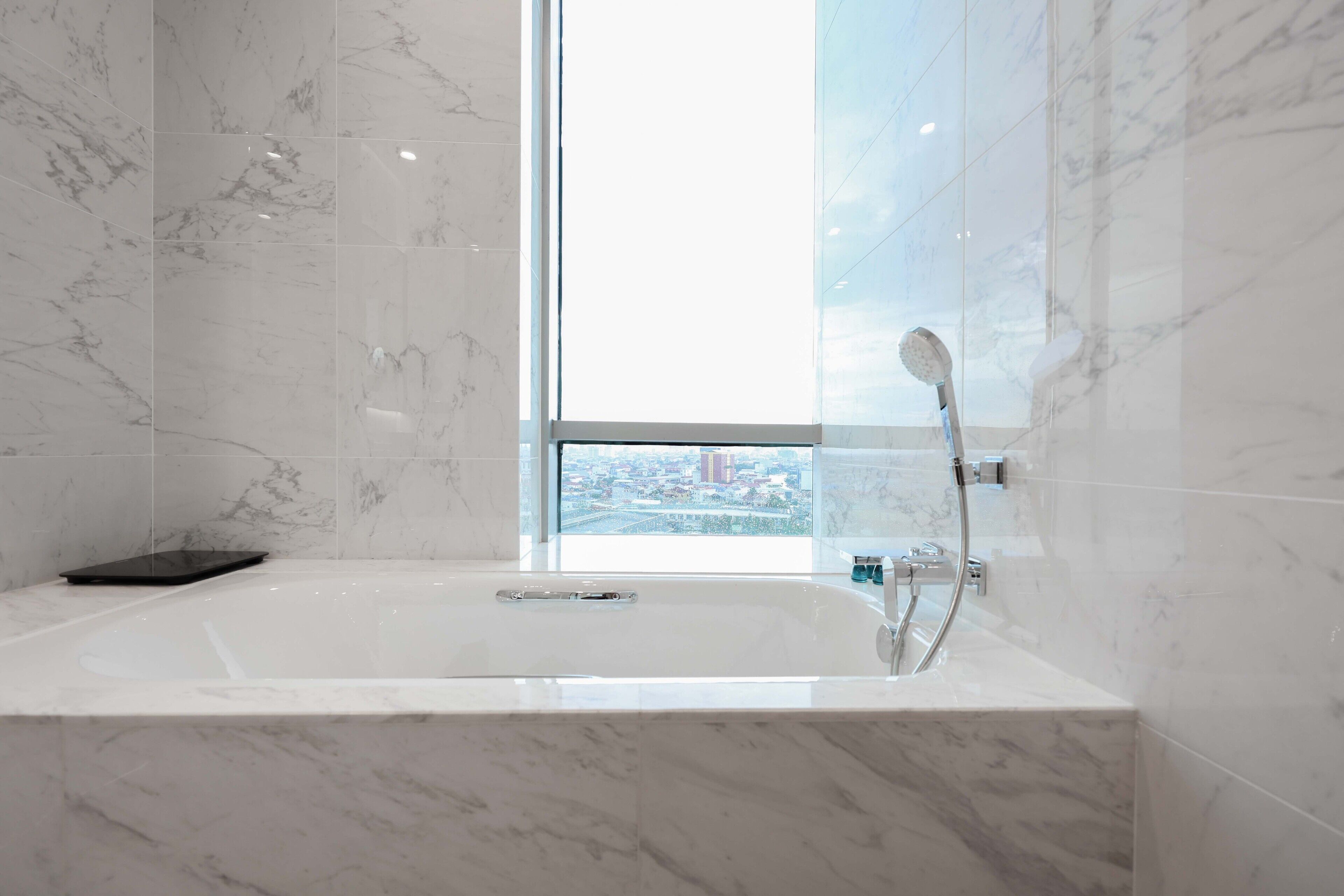 Une salle de bain moderne dotée d’une grande baignoire, d’une douchette à main et offrant une vue sur le paysage urbain grâce à une grande fenêtre.