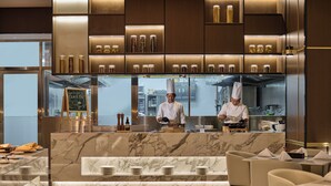Une cuisine moderne où des chefs préparent des plats, avec des plans de travail en marbre et un coin repas bien éclairé, équipé de chaises.