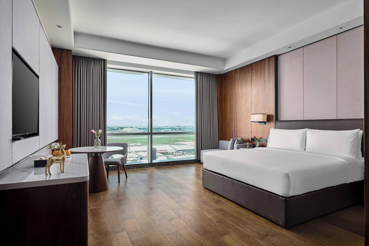 Ein modernes Hotelzimmer mit einem großen Bett, einem Schreibtisch mit Fernseher, einem Essbereich und Blick auf das Flughafengelände.