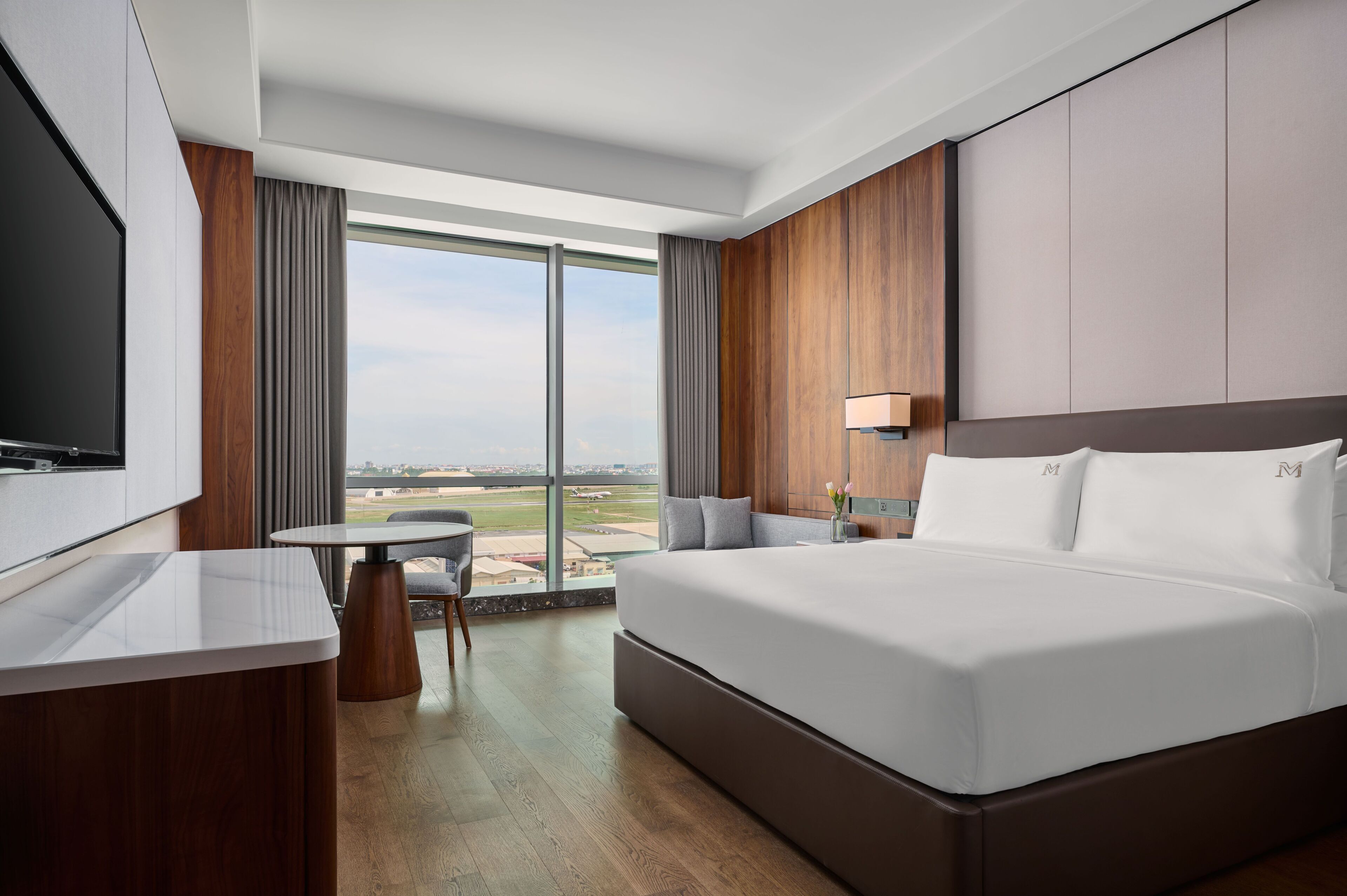 A modern hotel room with a large bed, a TV, a small table, and a view of the airport.