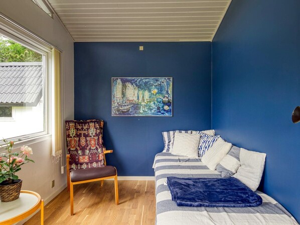A bedroom with a bed, a chair, a table with a plant, and a painting on the wall.