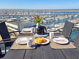 Una mesa de comedor con platos, una cafetera y un florero con flores, con vistas a un puerto deportivo con barcos.