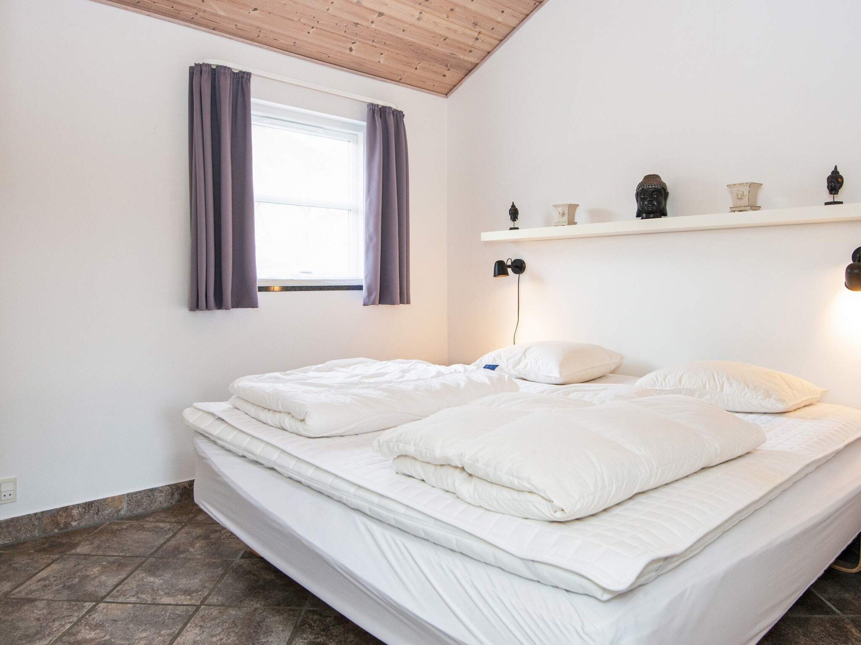 A bedroom with a bed, a window with curtains, a shelf with decorative items, and a wall-mounted lamp.