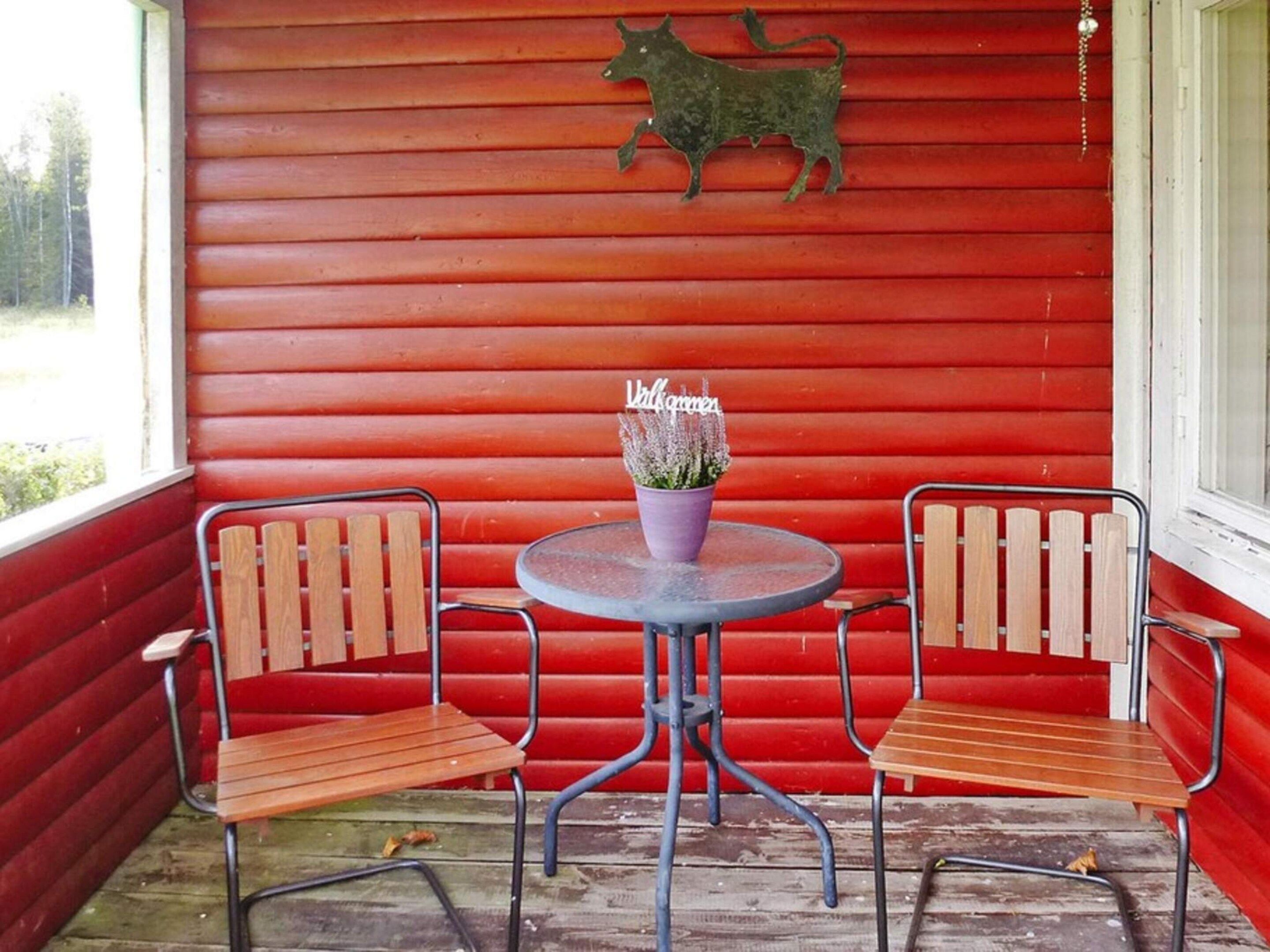 A red wooden porch with two wooden chairs and a round table, a cow decoration on the wall, and a potted plant on the table.