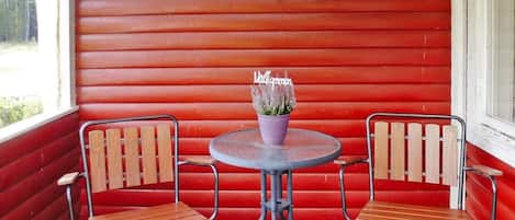 Une véranda en bois peinte en rouge, avec deux chaises en bois et une table ronde, une décoration représentant une vache sur le mur, et une plante en pot sur la table.