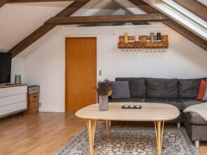 Een loft-achtige woonkamer met een houten plafond, een donkergrijze bank, een houten salontafel en een vloerkleed met een patroon.