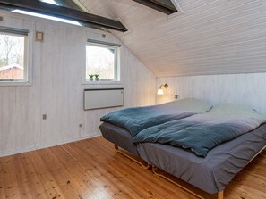 Een slaapkamer met schuine balken, houten vloer, een bed met grijze beddengoed, een raam met uitzicht op bomen en een wandradiator.
