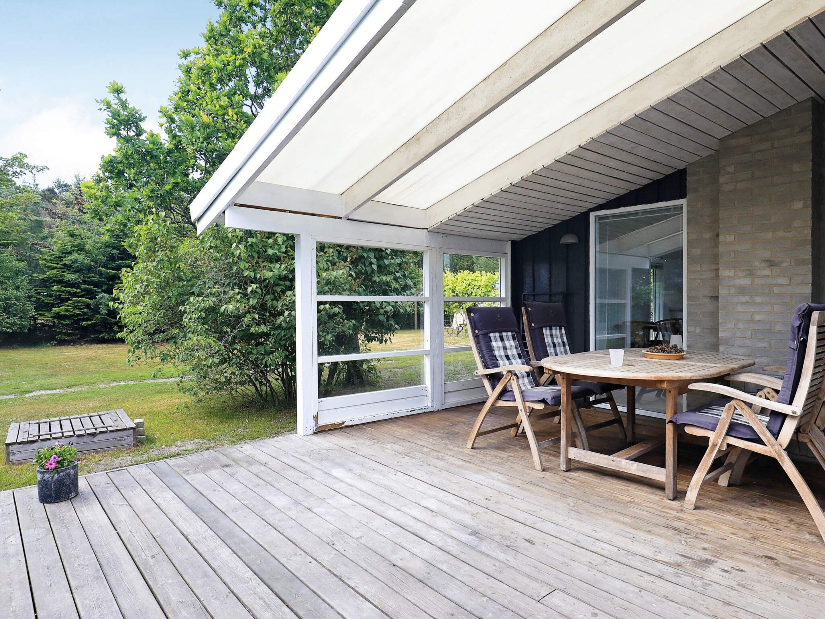 A covered outdoor seating area with a table and chairs, a wooden deck, and a view of the surrounding greenery.