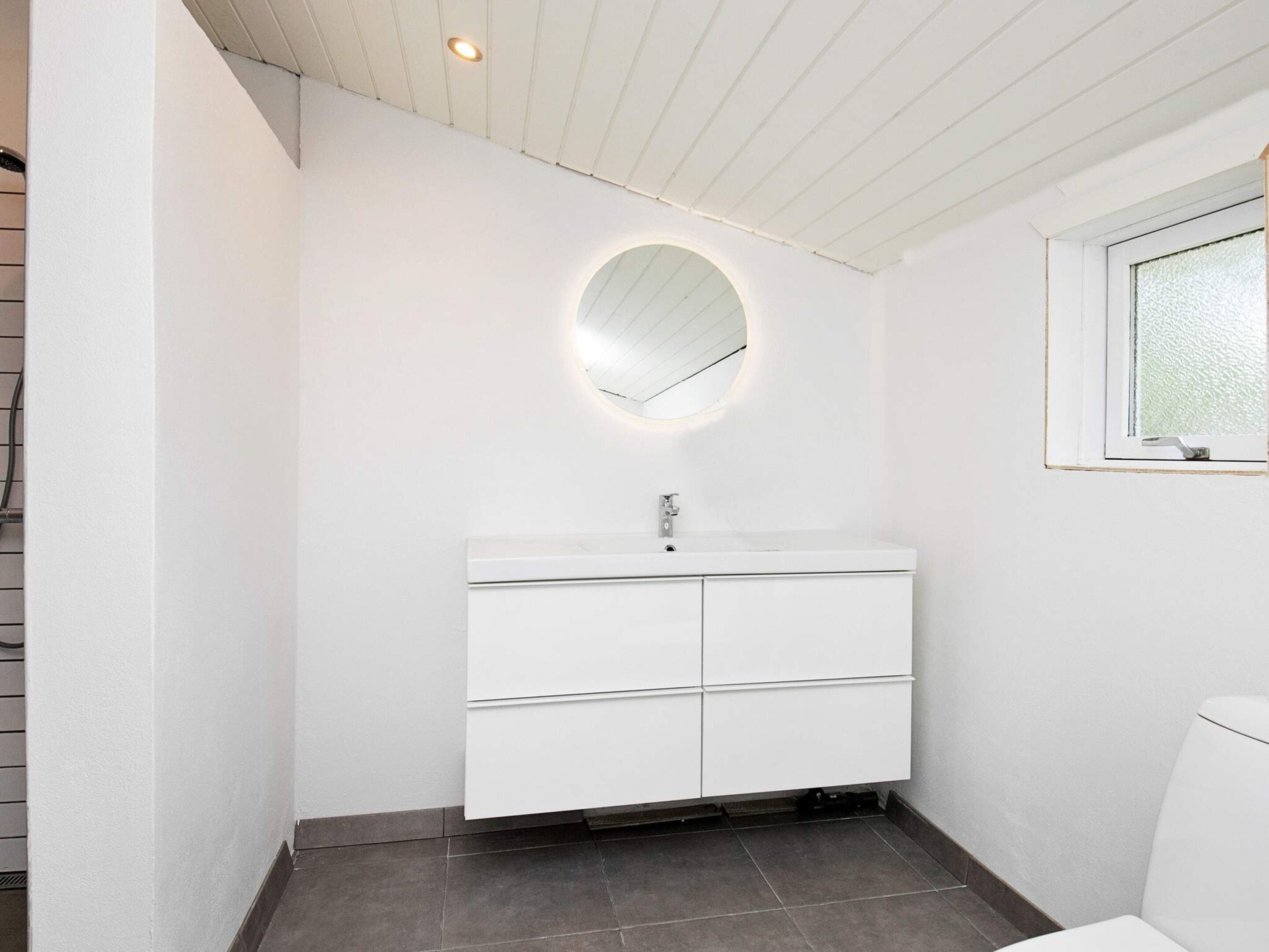 A modern bathroom with a white vanity, a round mirror, and a window.