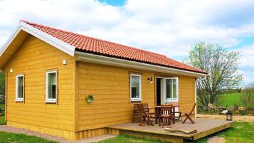 A yellow wooden house with a red-tiled roof, a covered patio with wooden furniture, and a grassy yard.