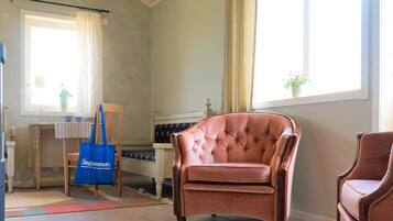 A room with a large window, a tufted leather armchair, a wooden chair, a small table with a striped tablecloth, and a blue tote bag.