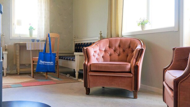 A room with a large window, a tufted leather armchair, a wooden chair, a small table with a striped tablecloth, and a blue tote bag. - 4 Star Holiday Home in Sandhem (Sandhem)