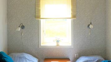 A room with two beds, a window with blinds, two wall-mounted lamps, and a small wooden table.
