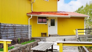 A yellow building with a red roof, a front door, and a small window. There is a wooden deck with a bench and a stone wall. - 9 Person Holiday Home in Fiskebackskil (Fiskebackskil)