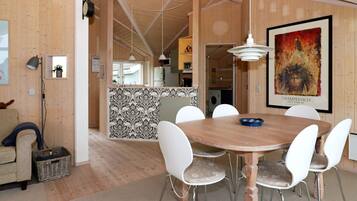 A dining area with a round wooden table and white chairs, a kitchen in the background, and a framed picture on the wall.